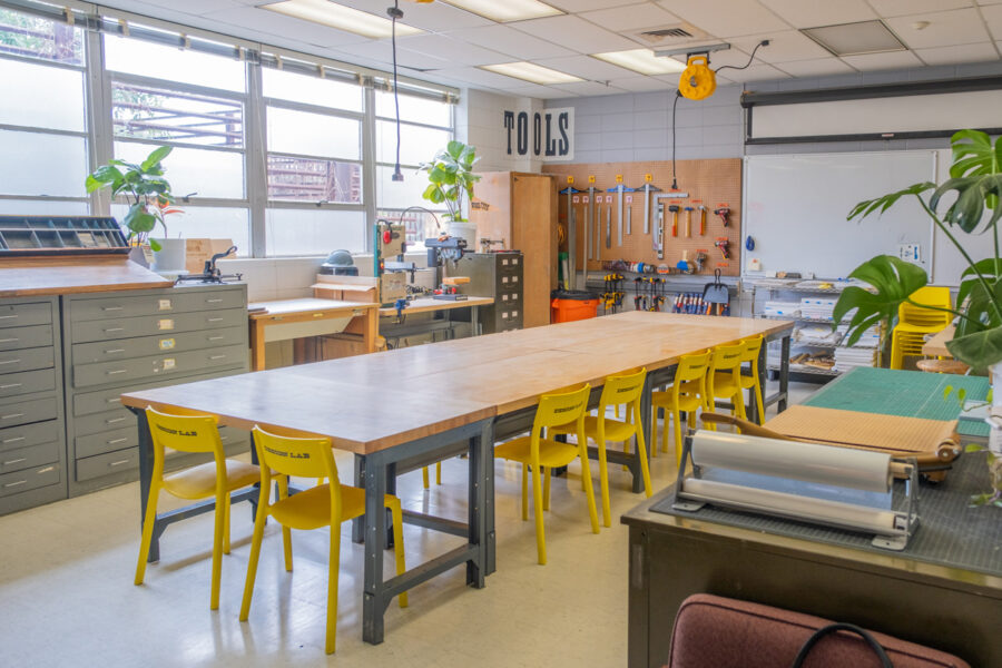 The wooden work tables in the Design Lab, with tools and equipment seen behind on a pegboard and against the windows. Plants and natural light fill the space