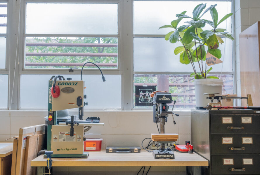 A closeup of a small tabletop bandsaw and drill press, with windows and a plant behind.