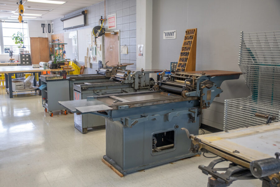 Looking across the shop at a row of various printing presses, made of painted gray metal.