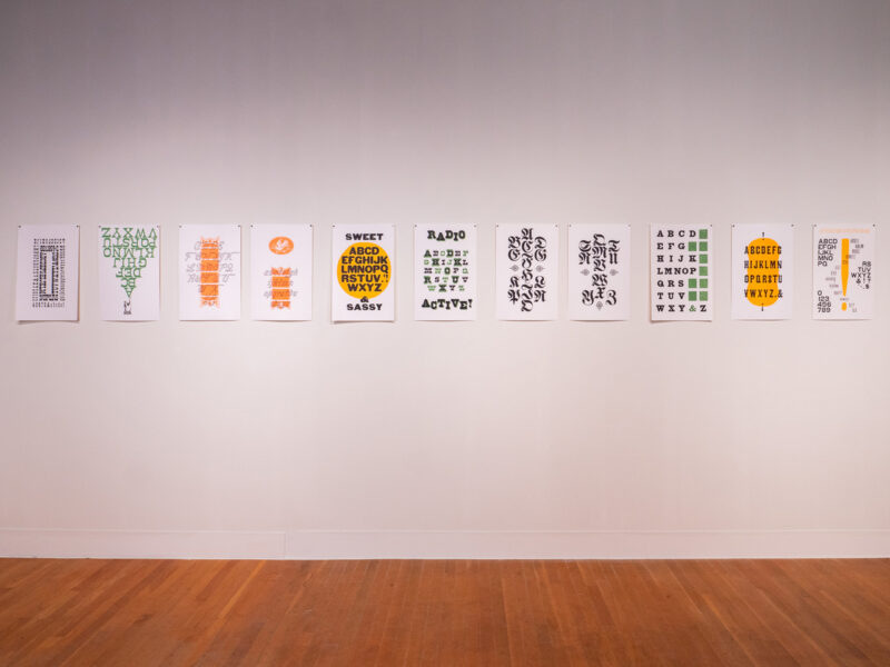 installation of RRK Folio Revisited Specimen prints on wall at the Visual Arts Center at UT Austin, eleven vertical prints in a row