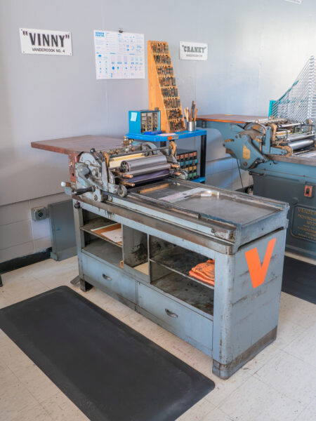 A cylinder printing press made of painted gray steel. Behind the press is our furniture and leading cart for setting type. The press has a white name plaque mounted to the gray wall above it, reading Vinny.