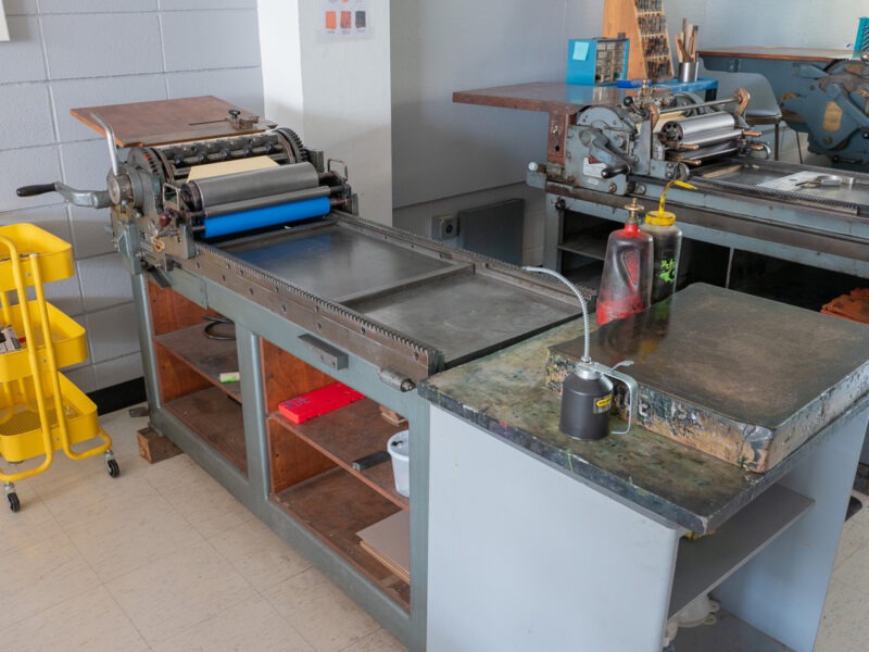 The Asbern press sits with a Vandercook No. 4 behind it. At the end of the press is an ink-mixing surface, with some oil and solvent containers. The press has a white name plaque mounted to the gray wall above it, reading Bernie.