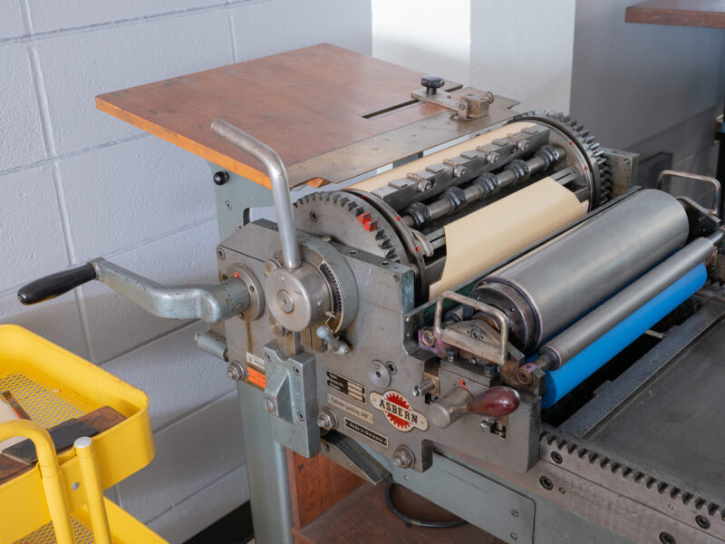 A closeup of the crank and cylinder of the Asbern press. This press has many wood and brass features, along with an additional lever on the cylinder for adjusting pressure. The Asbern label has a red gear icon.