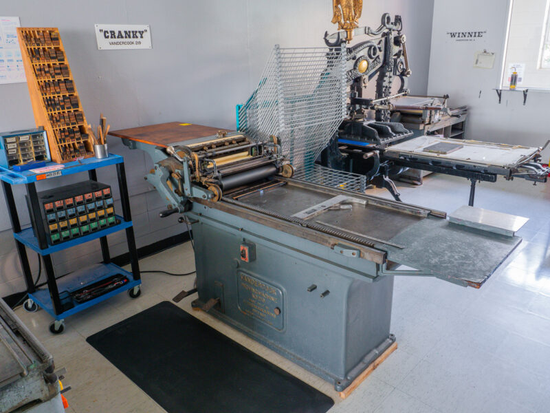 The Vandercook 219 OS, the base is made of painted gray cast iron. It prints up to 18 by 24 inch images. The Columbian platen press is seen behind the 219, illuminated by windows further back. The press has a white name plaque mounted to the gray wall above it, reading Cranky.