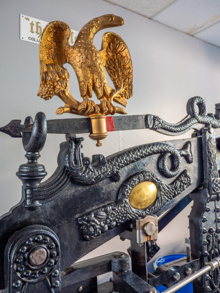 A closeup of the Columbian press, the brass and gold paint catches the light from the windows beyond. The press has a white name plaque mounted to the gray wall above it, reading The Eagle.