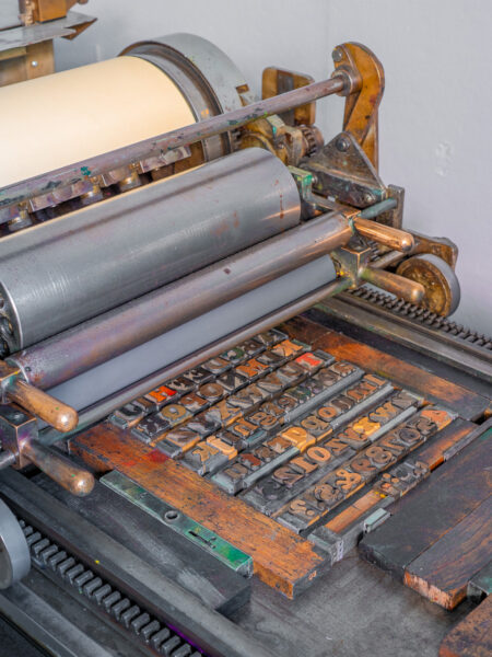 A closeup of the Vandercook No. 4 pressbed, there’s colorful wood type setup on the press, secured with furniture and leading. The cylinder of the press rolls above the type.