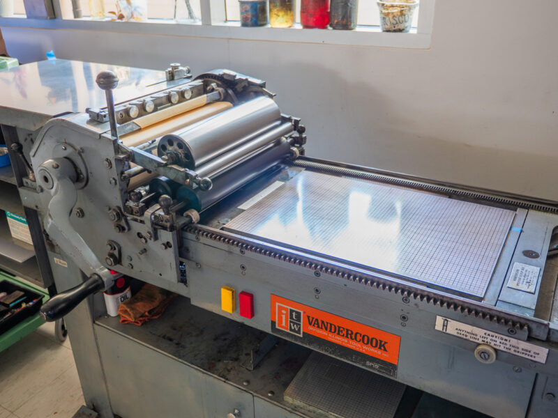 The SP15 glistens in the sunlight next to the window. The Vandercook label is a large orange rectangle below the pressbed. Containers for oil and solvent sit on the window sill beyond.