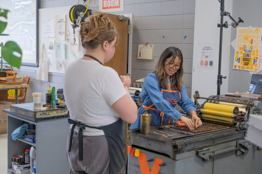 A student cleaning the Vandercook No. 4 printing press, with a wood type lockup on the pressbed. The rollers have yellow ink on them. Another staff member stands in the foreground.