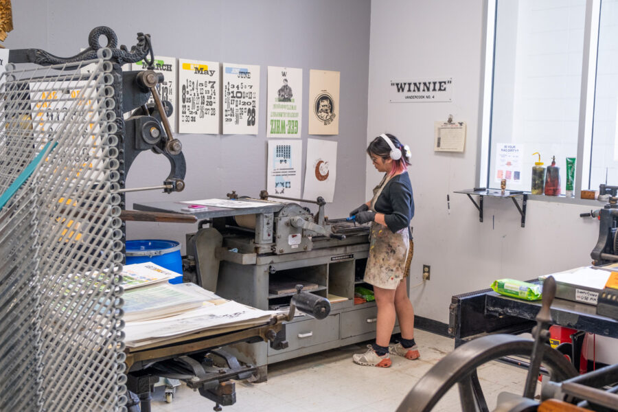 A student working on a lockup on Winnie, the Vandercook No. 4 printing press. Colorful prints hang on the wall behind