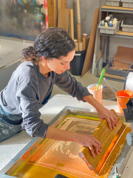 A student in the process of screen printing a poster, they hold a large squeegee and pull orange ink across the screen