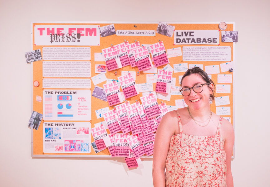 A student in front of their project installed at the Visual Art Center. The project is titled The Fem Press, and features pink and black booklet publications pinned to a corkboard.