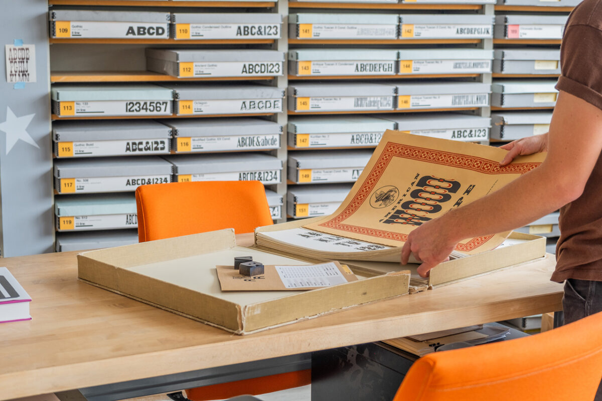 the large clamshell box from American Wood Type 1828-1900, Volume I, open on the wood table in the Design Lab office, in front of the wall of colorful type boxes. A person is handling the cover page, leafing through the sheets.