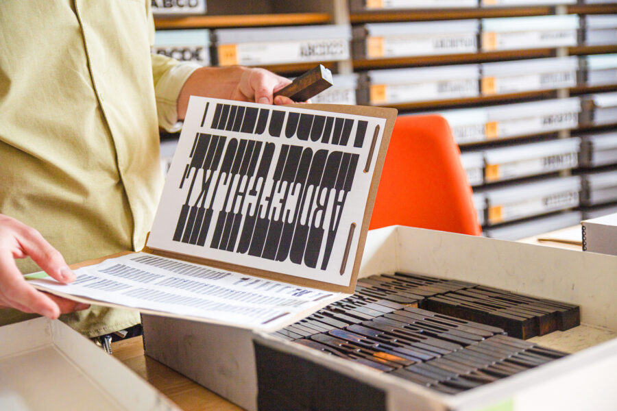 hands holding an open folder that holds a digitally printed wood type specimen example atop a box holding the skinny wood type blocks, one of the tallest in the collection called No. 117